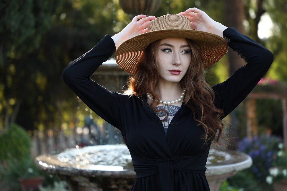 Natasha Rossi in pearls, a sun hat, and a black long sleeve v-neck top.